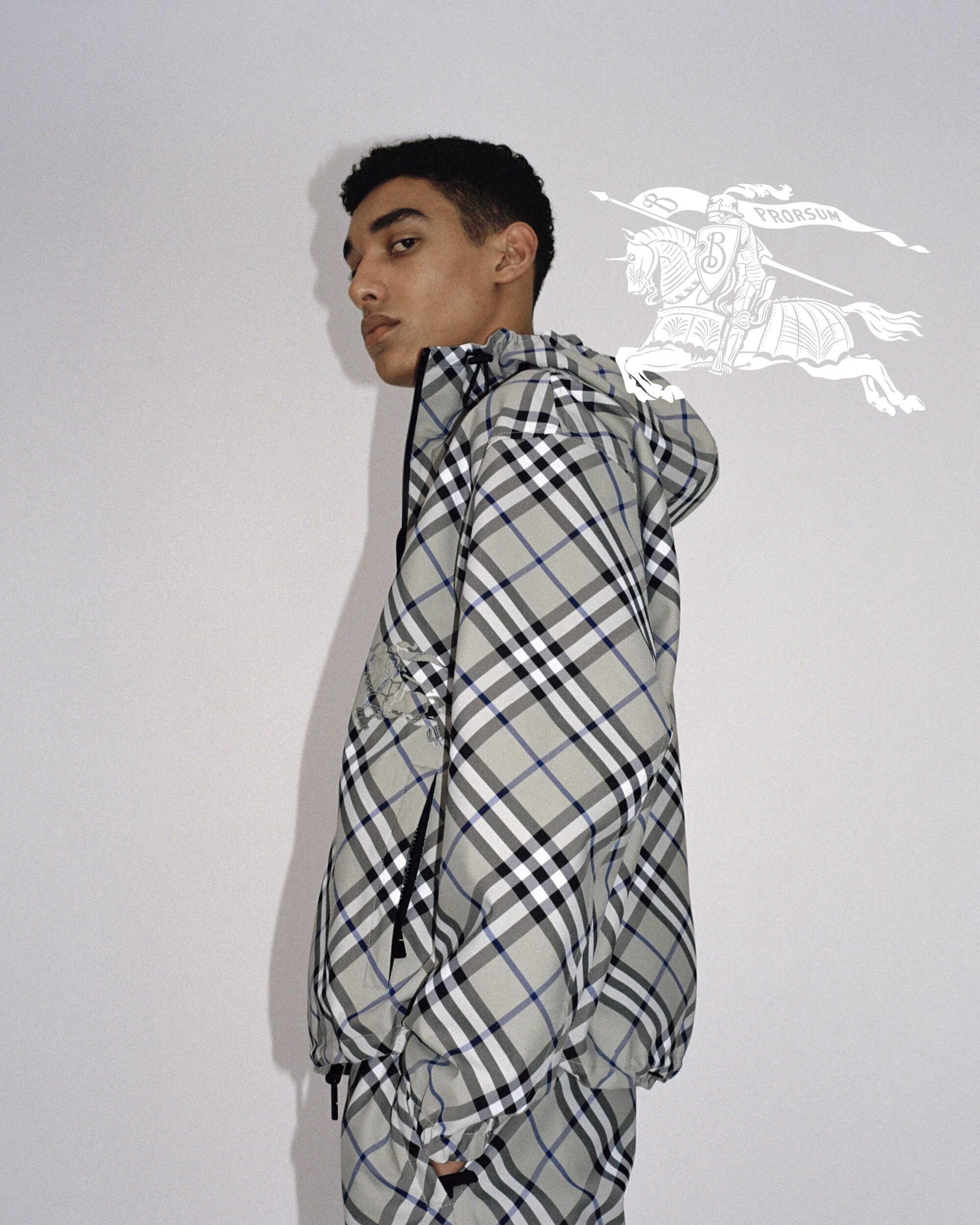 Adult male fashion model poses indoors wearing a grey plaid hooded jacket in a studio setting against a grey backdrop.