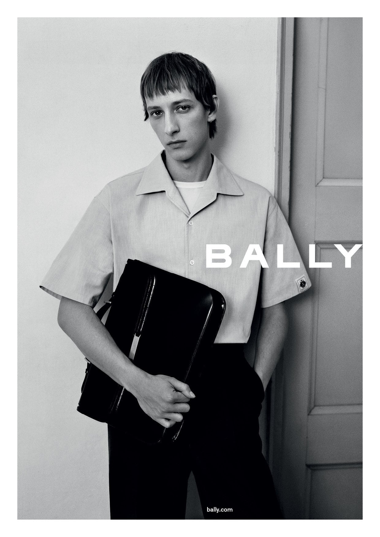 Fashion shot: Man in light button-down shirt holds black briefcase in a studio setting, showcasing modern apparel and accessories.