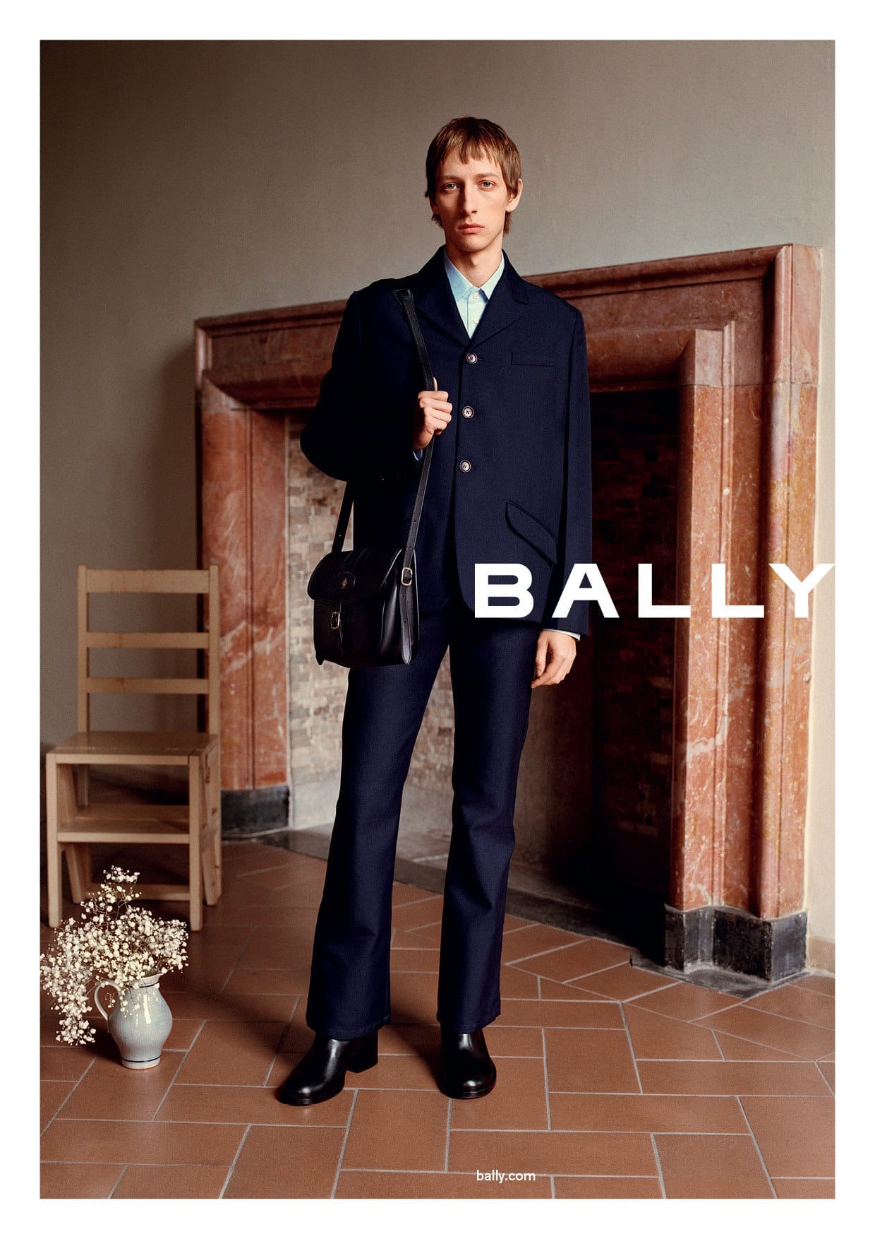 Bally fashion advertisement features a slender male model in a navy suit, shoulder bag, and black leather boots posing indoors.