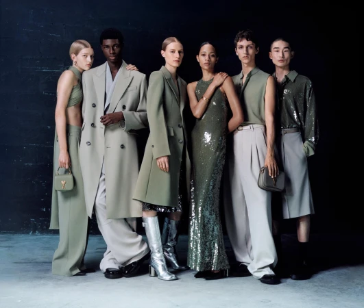 A group of six diverse fashion models showcase a collection of green and neutral-toned apparel against a dark backdrop, captured in a studio setting.