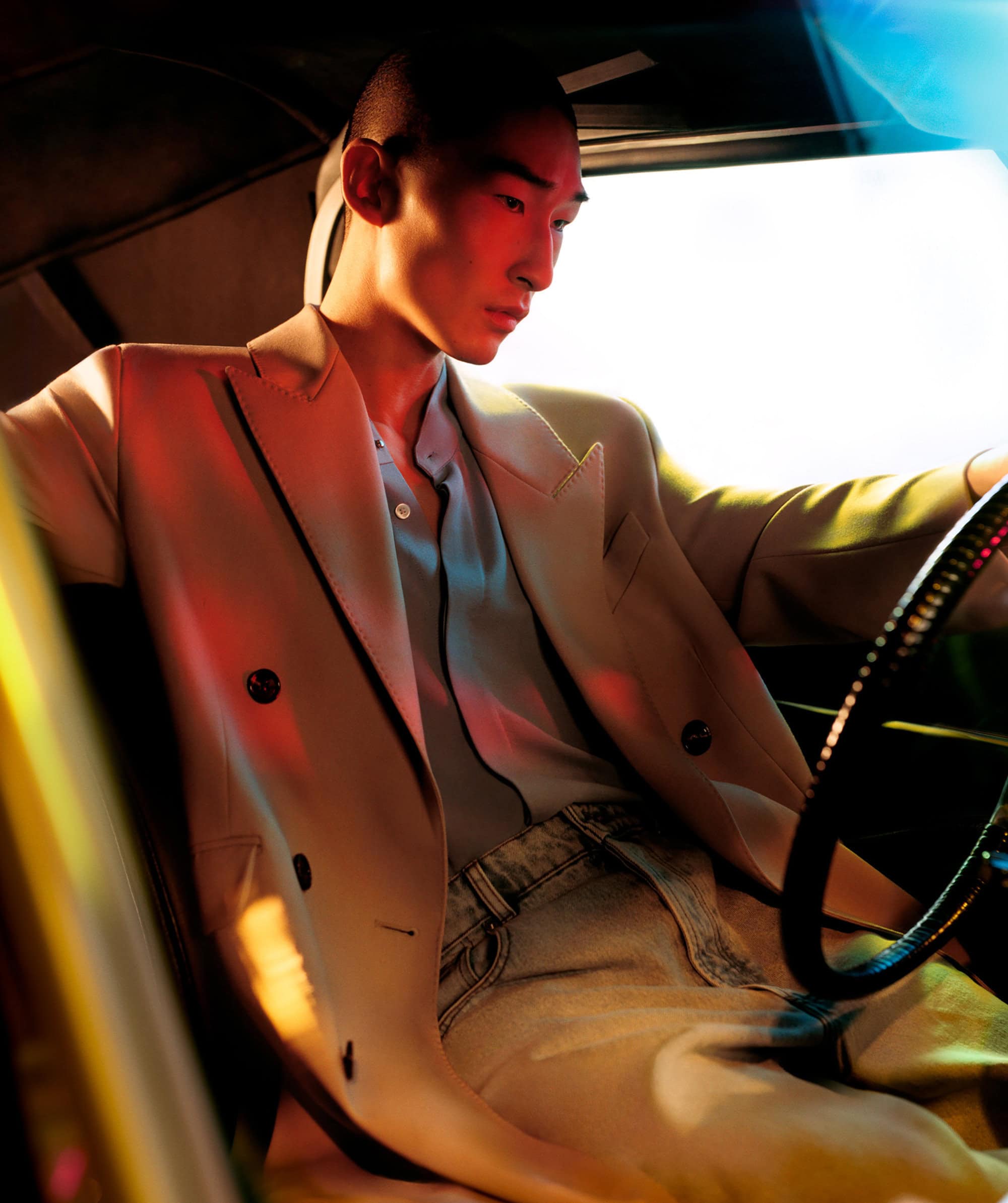 An Asian male model in a beige suit sits inside a car with a steering wheel, with red and blue lighting illuminating the scene.