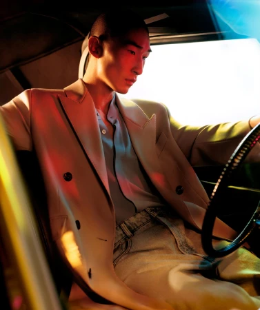An Asian male model in a beige suit sits inside a car with a steering wheel, with red and blue lighting illuminating the scene.