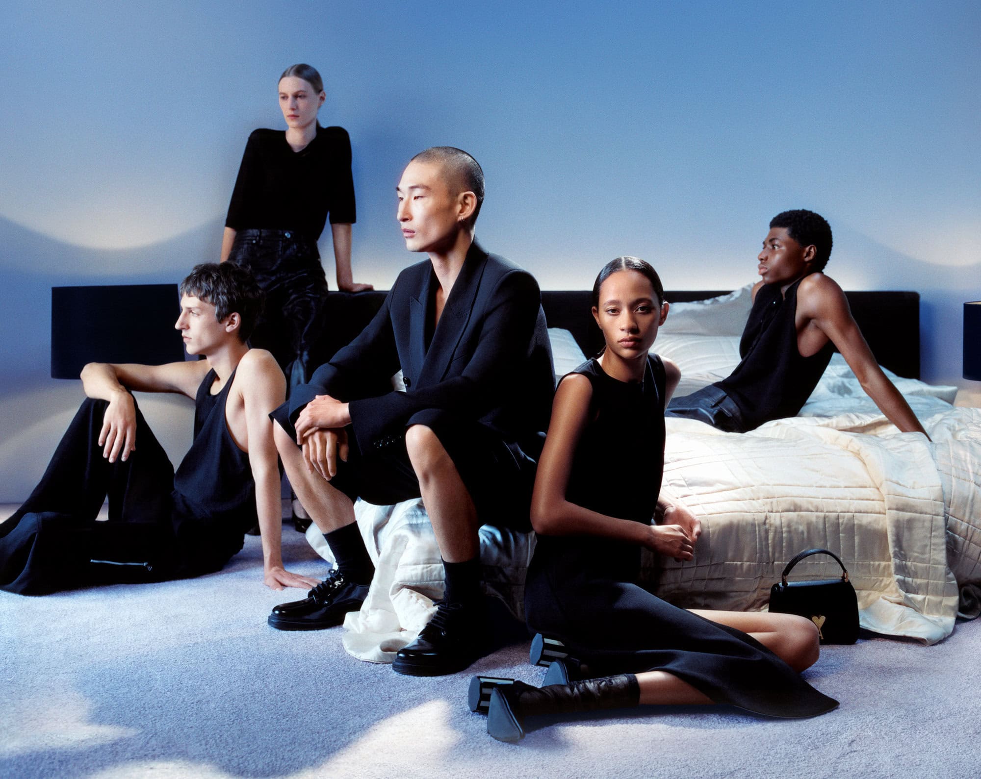 Editorial fashion shot of five diverse adult models in black clothing posing indoors on a bed with a blue background and neutral tones.