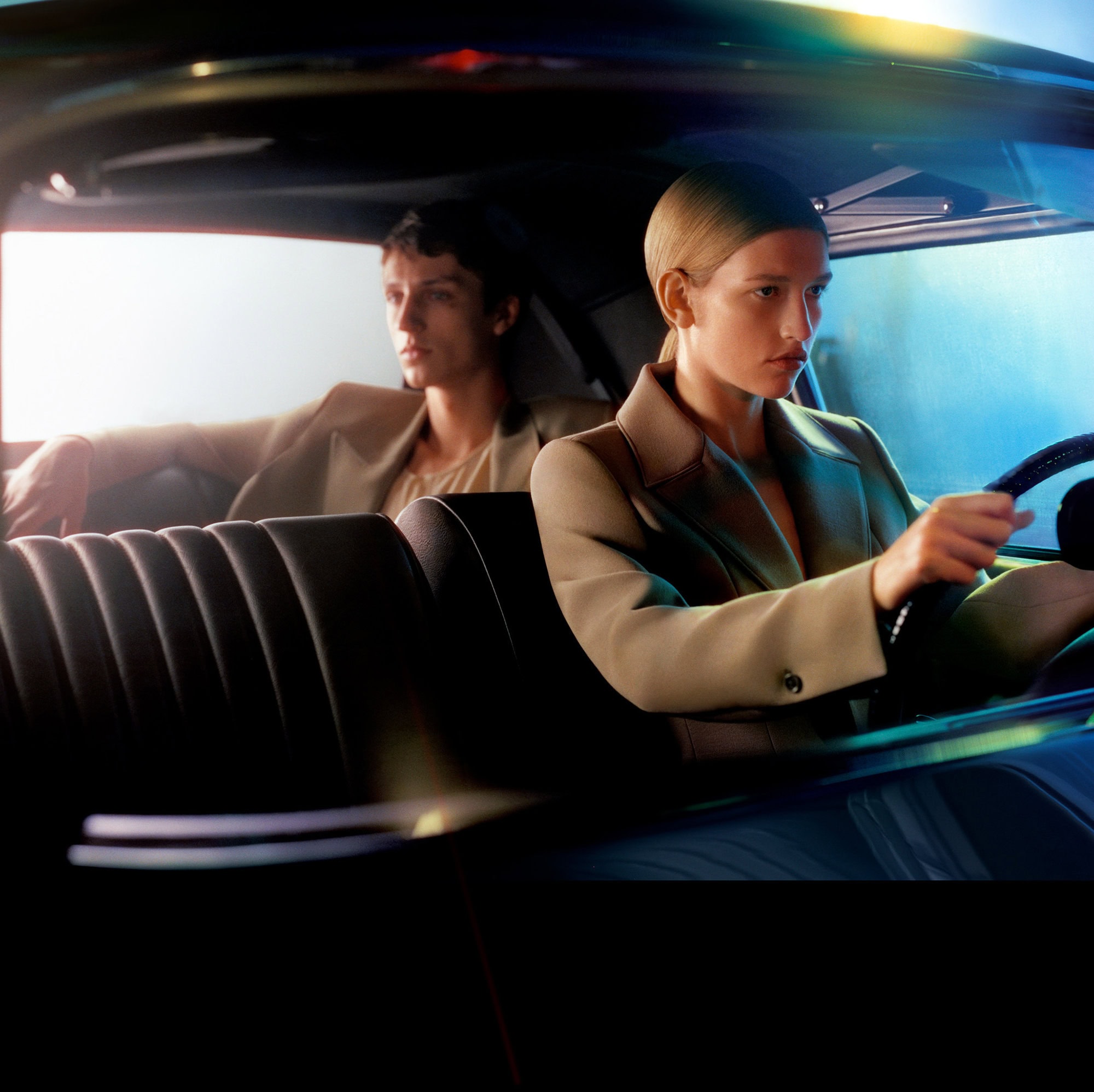 Couple in car: Blonde woman driving, man in passenger seat. Both are wearing neutral blazers and looking ahead. Dark interior.