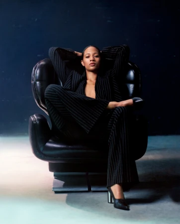 Elegant woman in pinstripe suit poses in a black chair against a dark backdrop. The portrait showcases fashion and design.