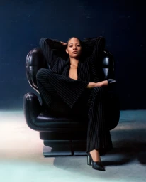 Model seated in a black armchair wearing a pinstripe pantsuit with hands behind head for Ami Paris