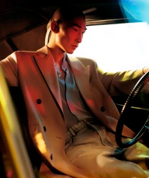 Man seated inside a car wearing a tailored blazer and holding a steering wheel, illuminated by dramatic red and blue lighting.