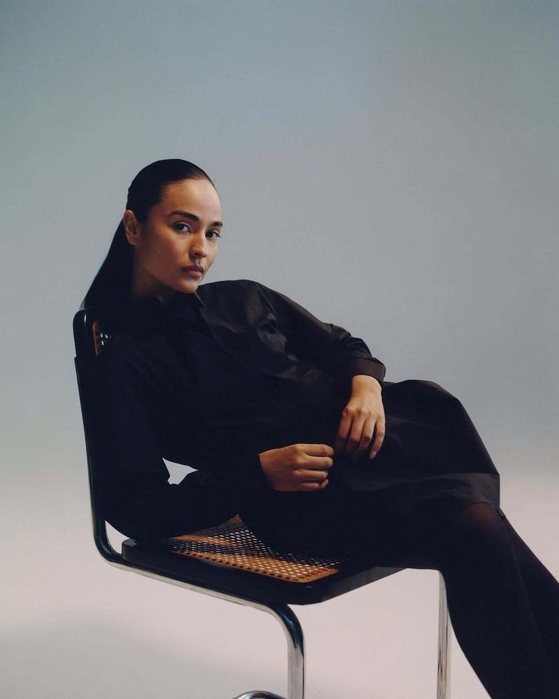 Portrait of a young woman in a black dress sitting in a chair with a light blue background looking at the camera in a studio.