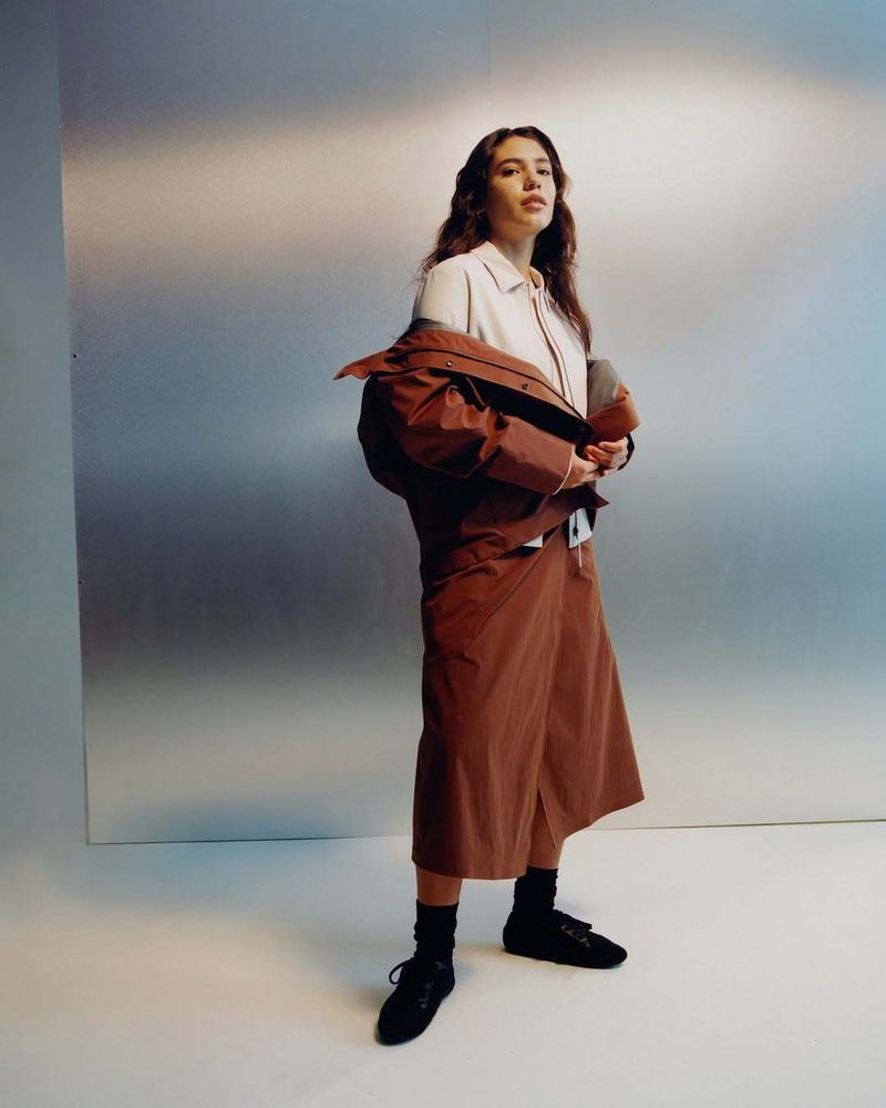 Fashion shot of a woman with long brown hair wearing a beige blouse, brown culottes, black socks, and black flats posing indoors.