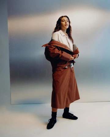 Fashion shot of a woman with long brown hair wearing a beige blouse, brown culottes, black socks, and black flats posing indoors.