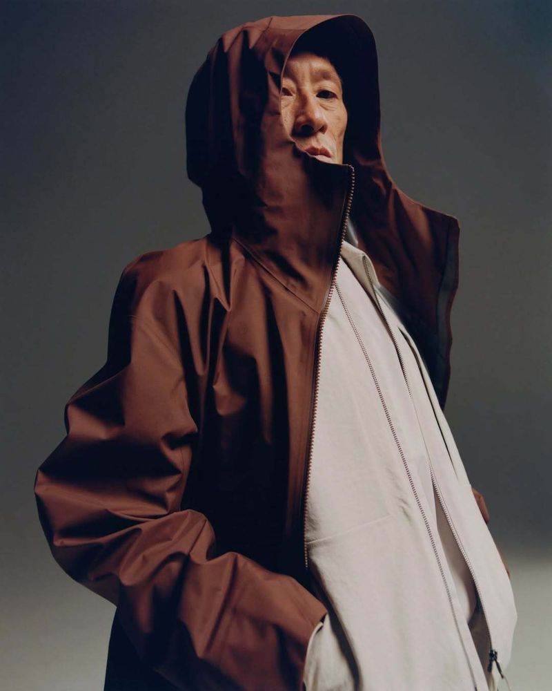 Fashion shot of an adult man in a brown hooded jacket against a gray studio backdrop, showcasing a blend of modern style and studio elegance.
