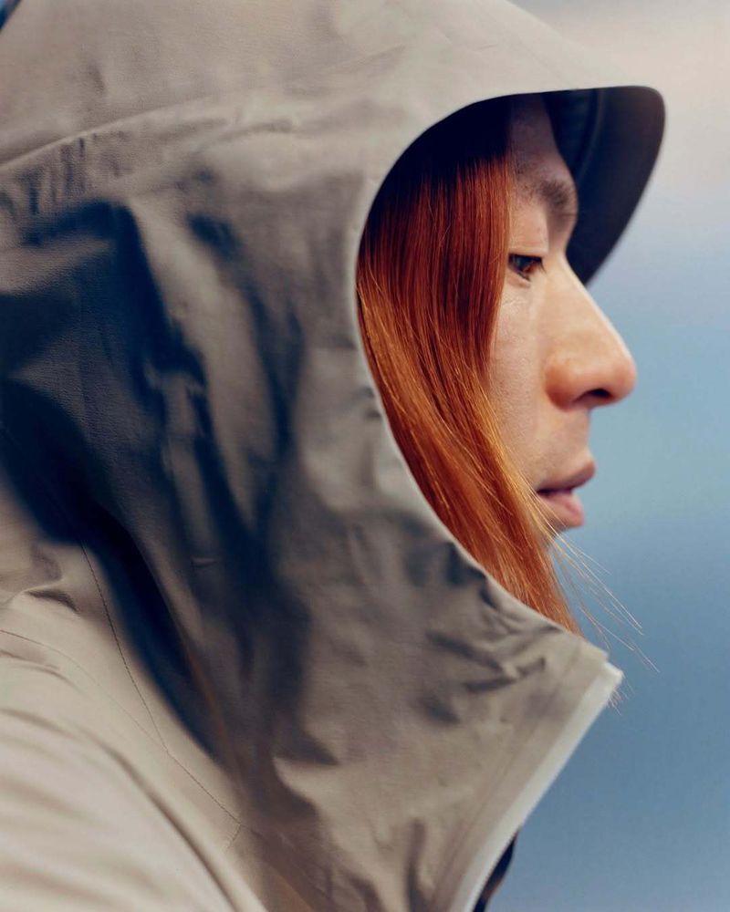 Close-up portrait of an adult with auburn hair wearing a grey hooded jacket, set against a blurred background of light blue and grey.