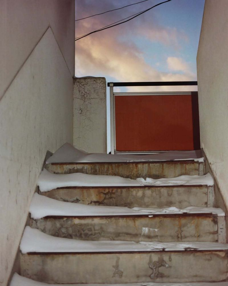 Snow-covered stairs lead to a rusty orange gate against a pastel sky in an outdoor urban setting. Concrete steps and wall, gate, sky.
