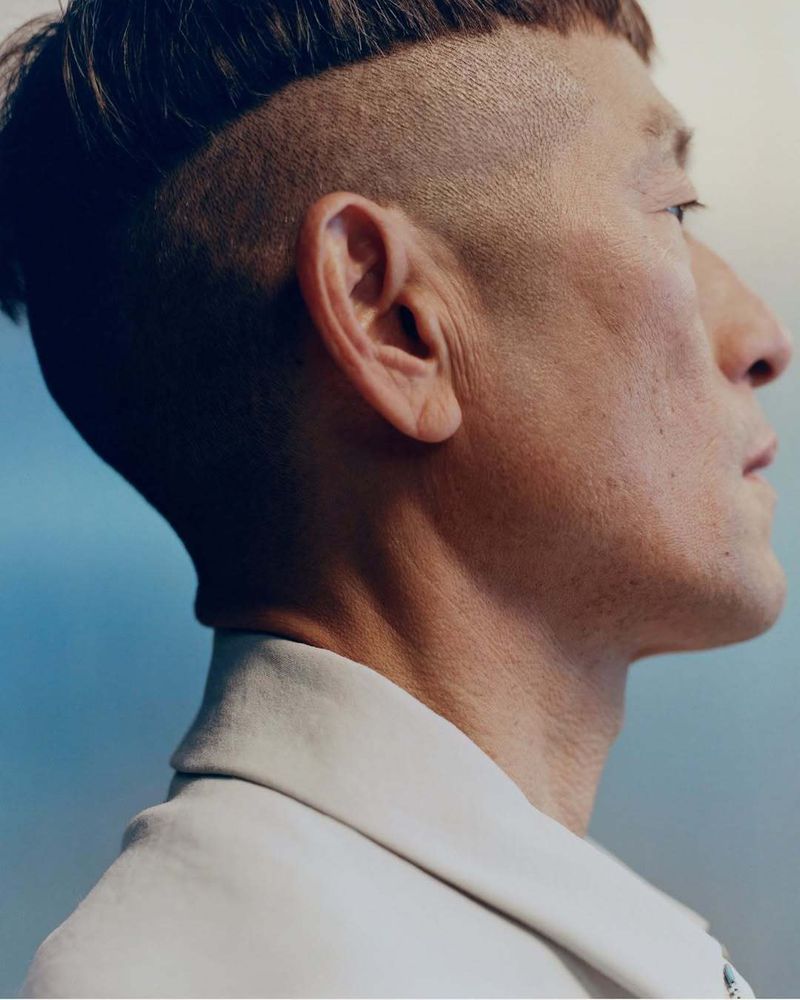Portrait of adult male with shaved head and fringe wearing a cream-colored jacket against a gradient blue and grey background.