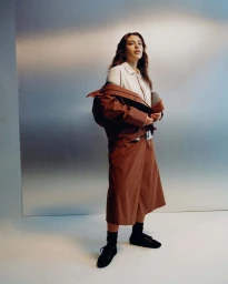 Model wearing a brown technical jacket and skirt set over a white shirt in front of a metallic wall.