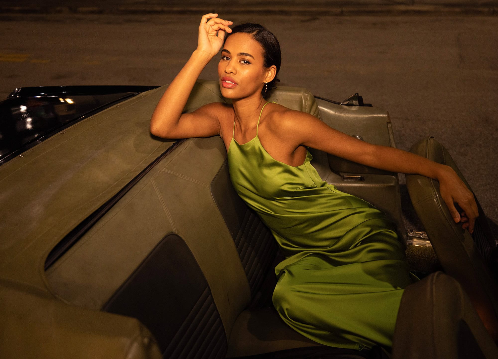 Elegant woman in a green dress poses inside a classic car at night with soft lighting highlighting her features and the car's interior.