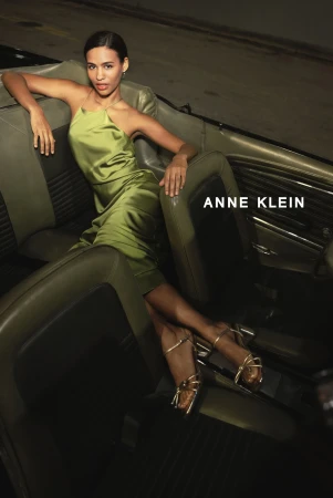 Elegant adult woman in a satin dress reclines in a classic car, showcasing gold strappy heels, indoors with ANNE KLEIN text.