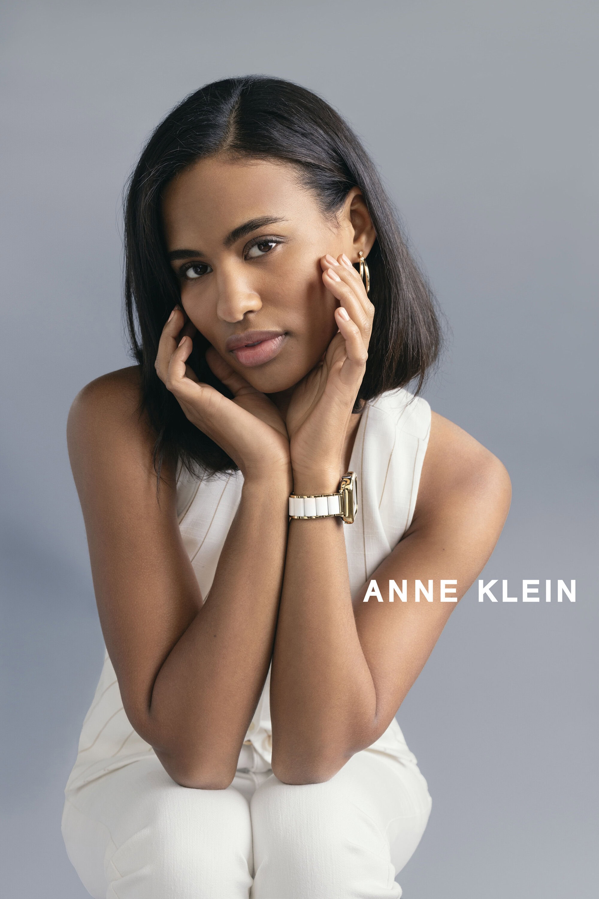 Woman in white sleeveless top with gold watch and earrings posing against a grey seamless studio background.