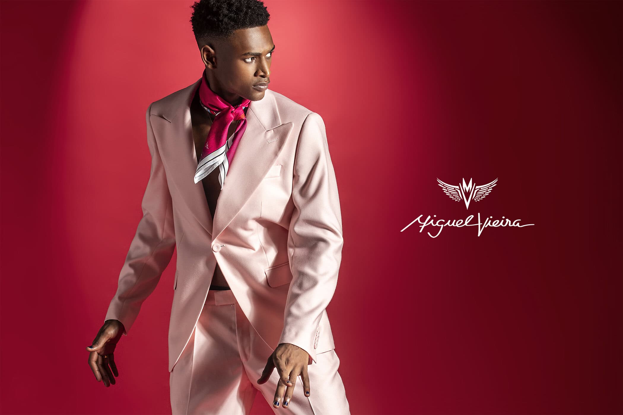 Fashionable young Black man in a pink suit and silk scarf posing in front of a red background with signature logo detail.