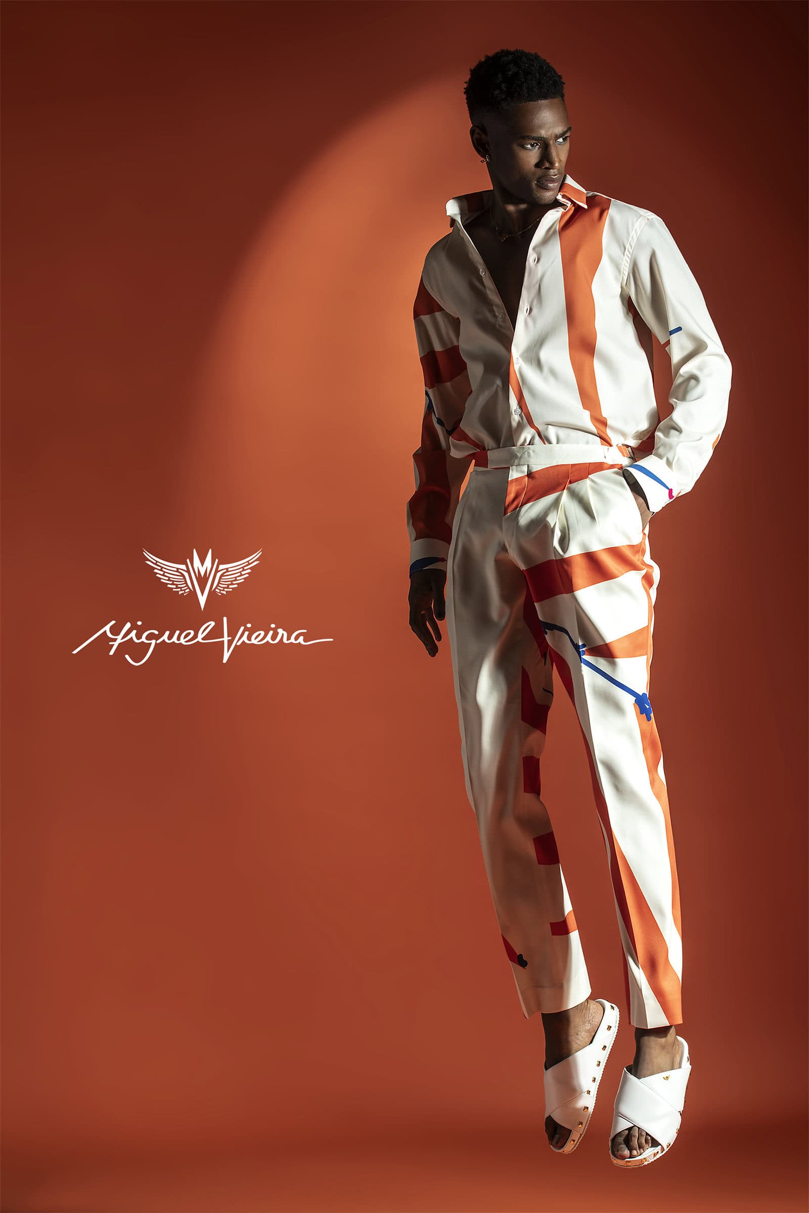 A well-lit studio fashion shot showcases a stylish adult male wearing a patterned shirt and pants, floating against an orange backdrop.