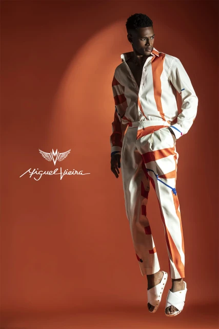 A well-lit studio fashion shot showcases a stylish adult male wearing a patterned shirt and pants, floating against an orange backdrop.