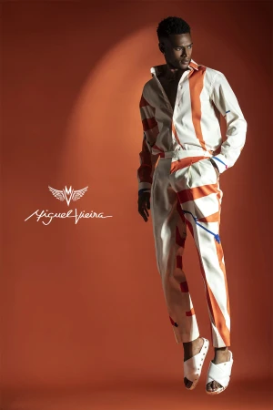 A well-lit studio fashion shot showcases a stylish adult male wearing a patterned shirt and pants, floating against an orange backdrop.