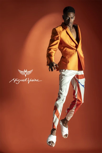 Fashionable African American man in orange blazer and white pants leaps in the air against a vibrant orange backdrop in a studio setting.