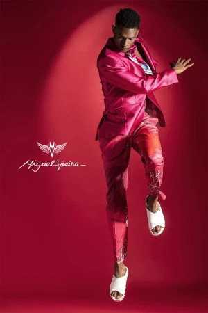 A man in a vibrant pink suit mid-air, illuminated against a red backdrop, showcasing fashion and style with a touch of performance.