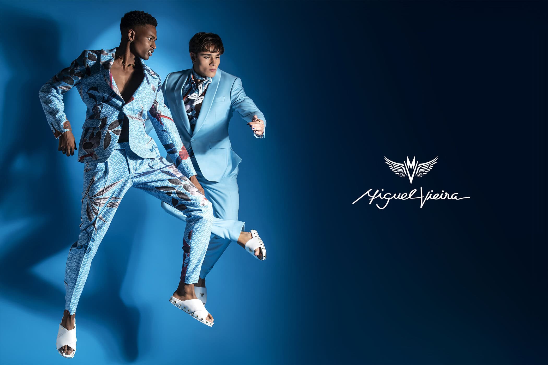Two men model light blue suits in a studio fashion shot, one in patterned suit, mid-air jump, against a solid blue backdrop.