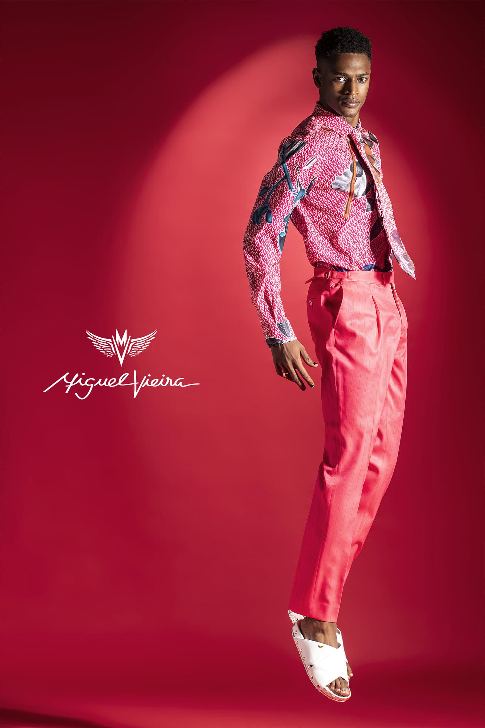 Full-shot of a Black male model in a patterned shirt, pink pants, and white sandals jumping in front of a red studio background.