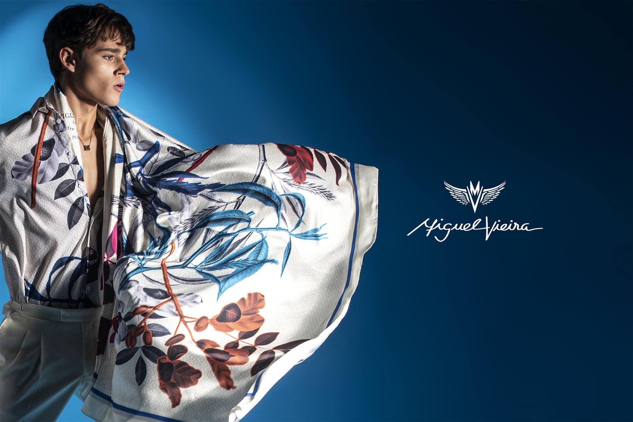 A young male model presents a floral silk scarf, casually draped over his shoulders, against a deep blue backdrop for a fashion campaign.