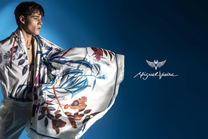 A young male model presents a floral silk scarf, casually draped over his shoulders, against a deep blue backdrop for a fashion campaign.