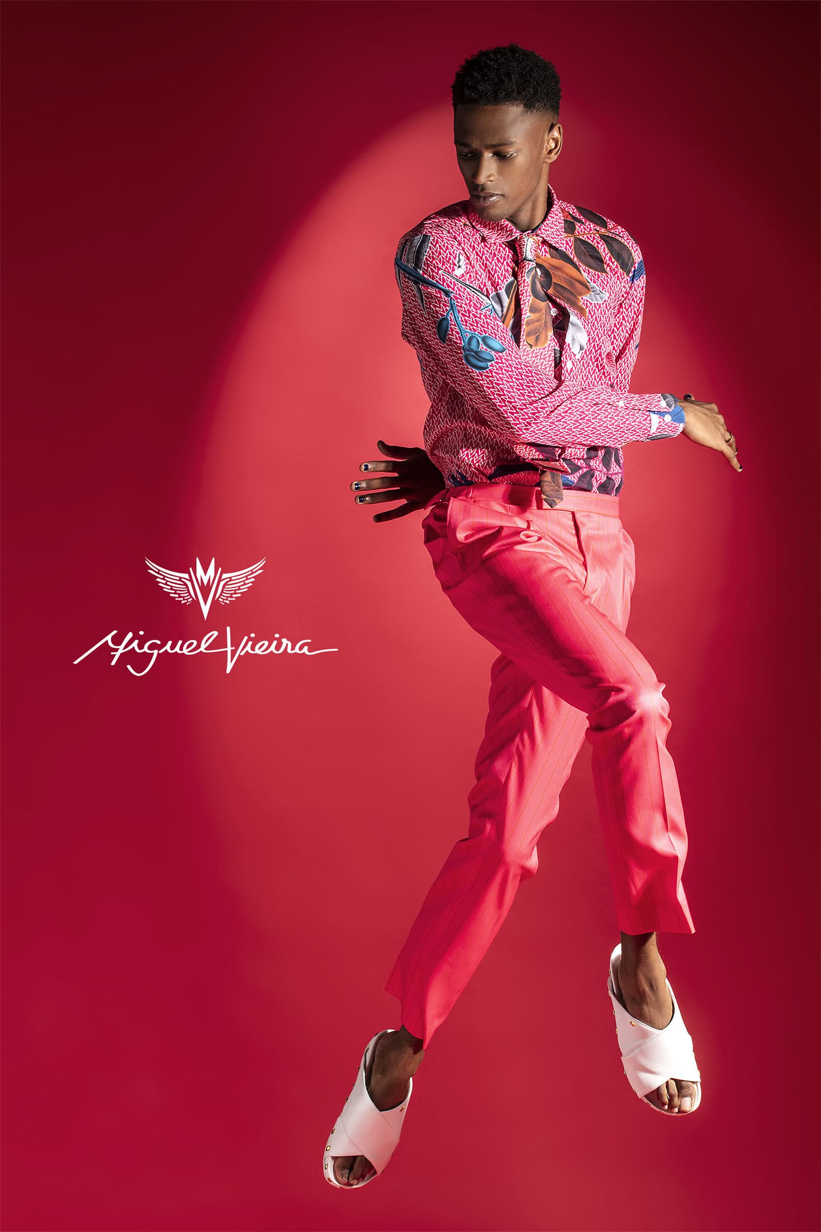 Young man in patterned shirt and red pants mid-air against a red backdrop, fashion portrait showcasing unique style, studio lighting.