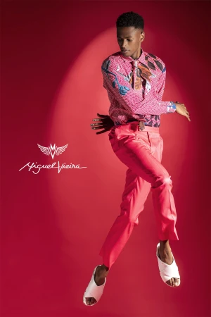 Young man in patterned shirt and red pants mid-air against a red backdrop, fashion portrait showcasing unique style, studio lighting.
