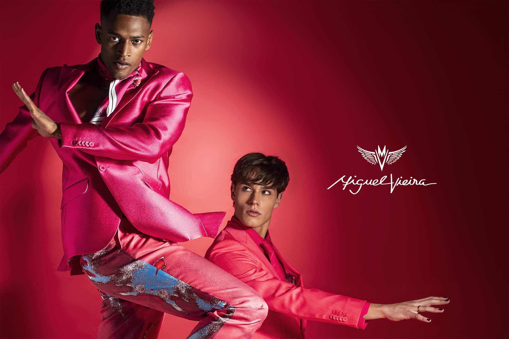 Two male models pose against a vibrant red background, showcasing a pink suit with blue and silver accents, complemented by a logo.
