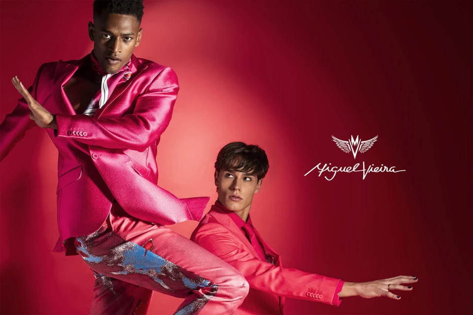 Two male models pose against a vibrant red background, showcasing a pink suit with blue and silver accents, complemented by a logo.
