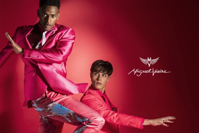 Two male models pose against a vibrant red background, showcasing a pink suit with blue and silver accents, complemented by a logo.