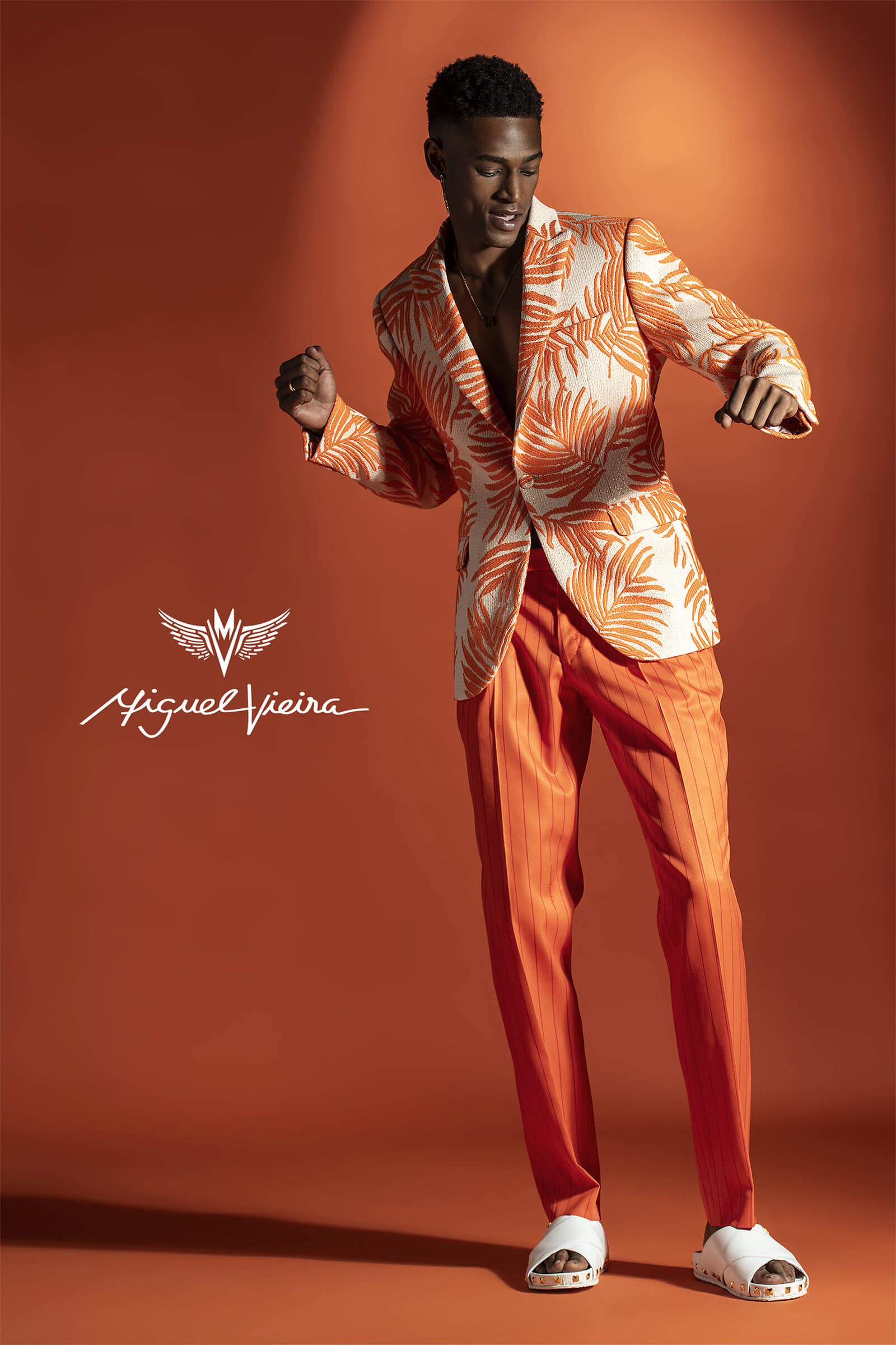 Male model poses in a patterned blazer and orange pants against a matching backdrop. He is captured mid-movement with a dynamic pose.