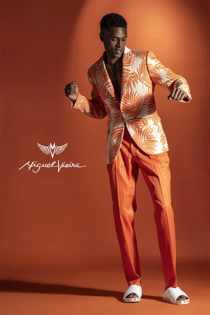 Male model poses in a patterned blazer and orange pants against a matching backdrop. He is captured mid-movement with a dynamic pose.