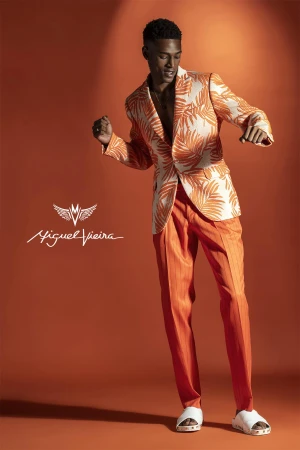 Male model poses in a patterned blazer and orange pants against a matching backdrop. He is captured mid-movement with a dynamic pose.