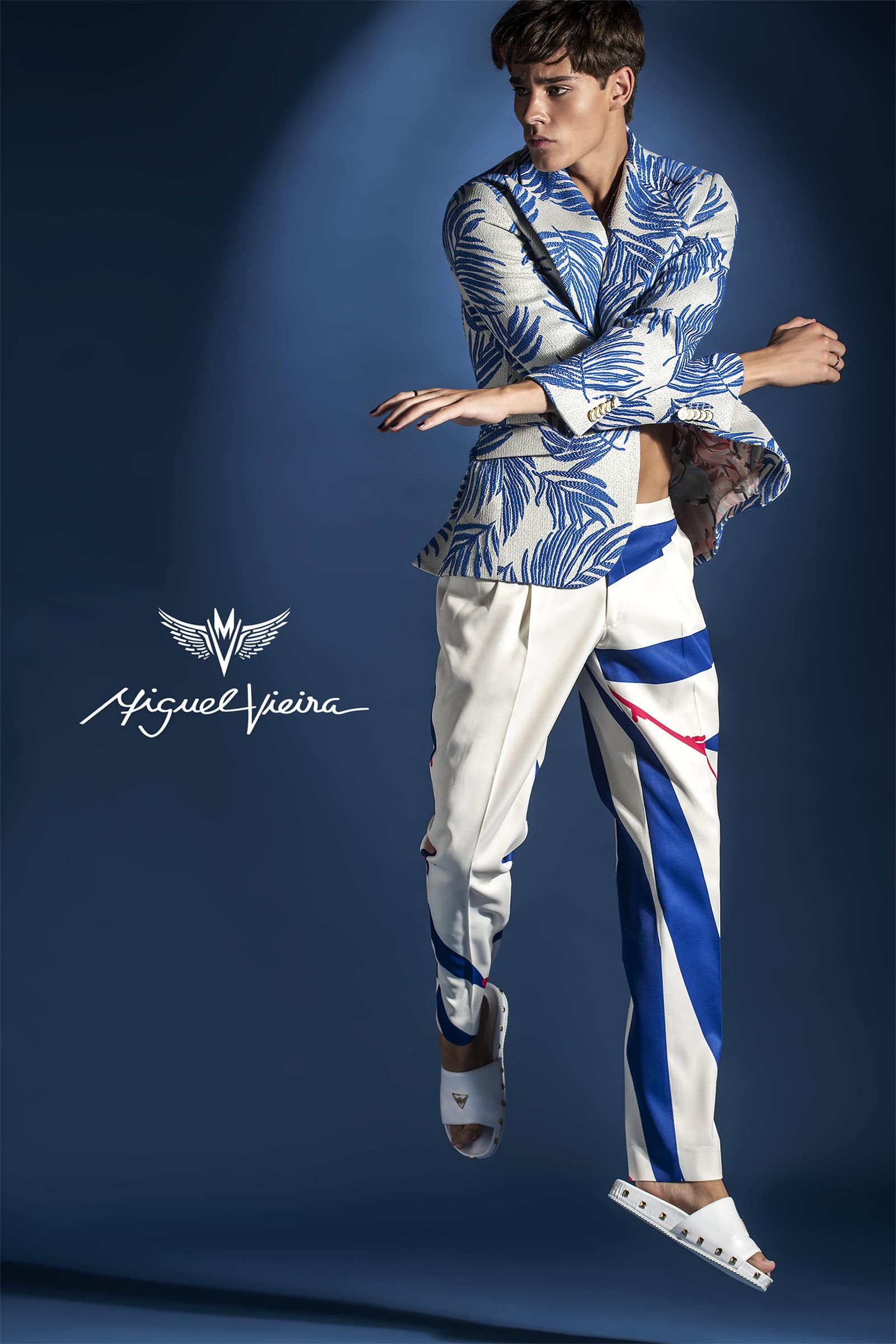 Fashionable male model levitates in a blue and white patterned jacket and white pants against a blue background in a studio fashion shot.