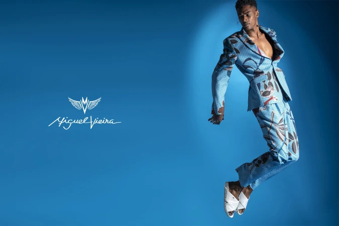 Adult male model in a patterned blue suit jacket and pants jumping in a blue photo studio, sandals, and a logo on the left.