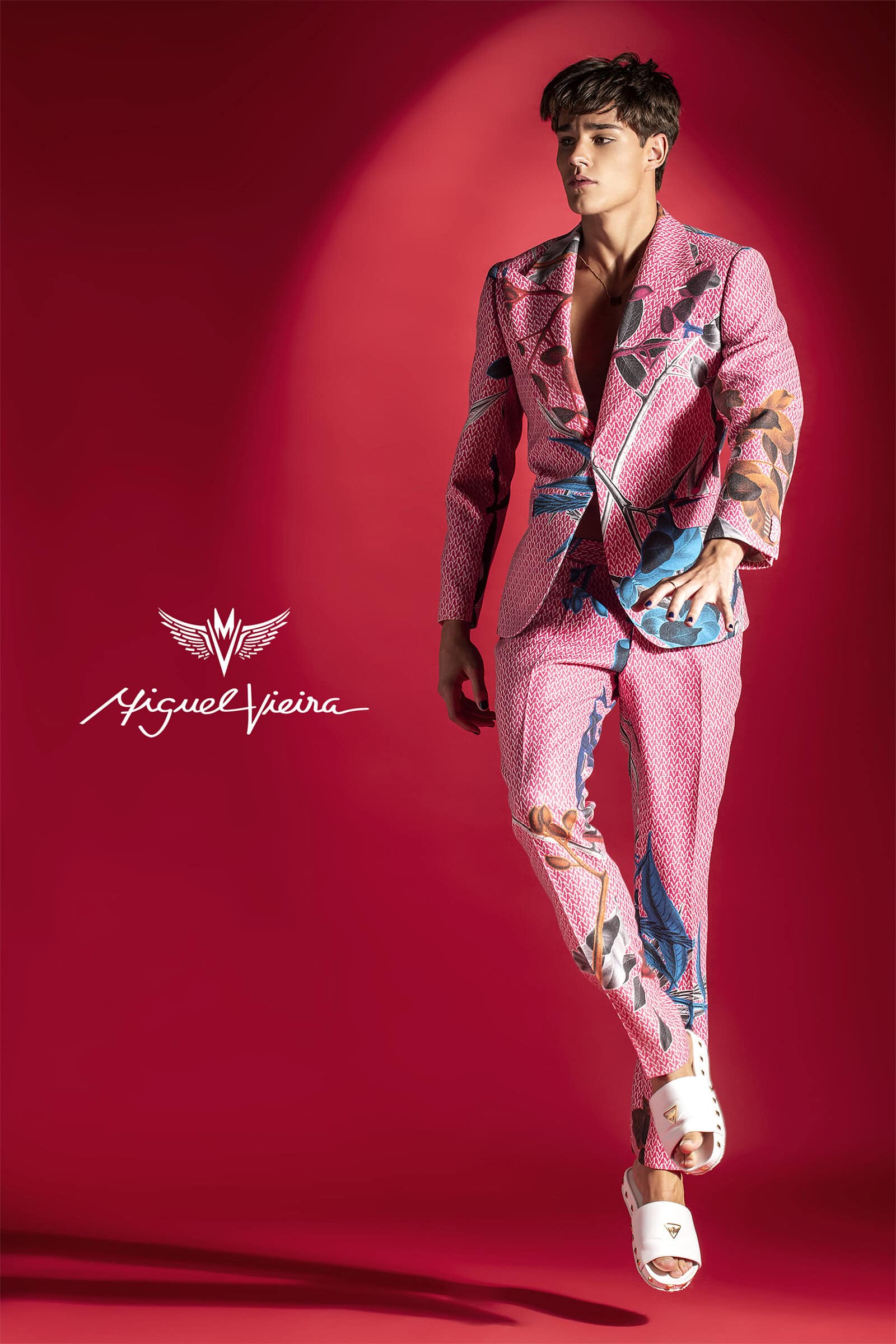 A young adult male model poses in a pink floral suit with white sandals against a red backdrop, showcasing a fashion-forward look.