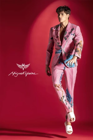 A young adult male model poses in a pink floral suit with white sandals against a red backdrop, showcasing a fashion-forward look.