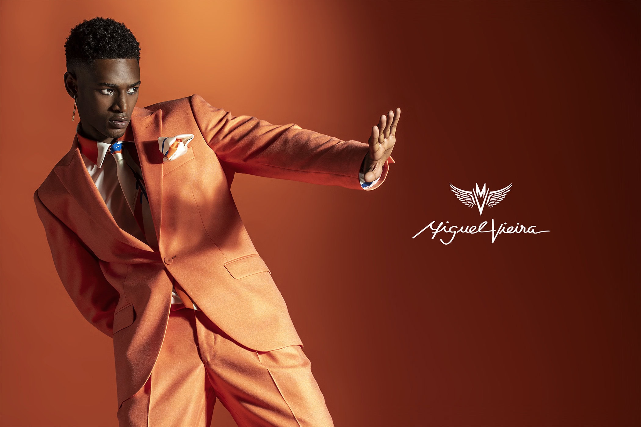 Stylish Black male model in a peach suit poses against a matching backdrop, showcasing modern fashion in a studio setting with logo.