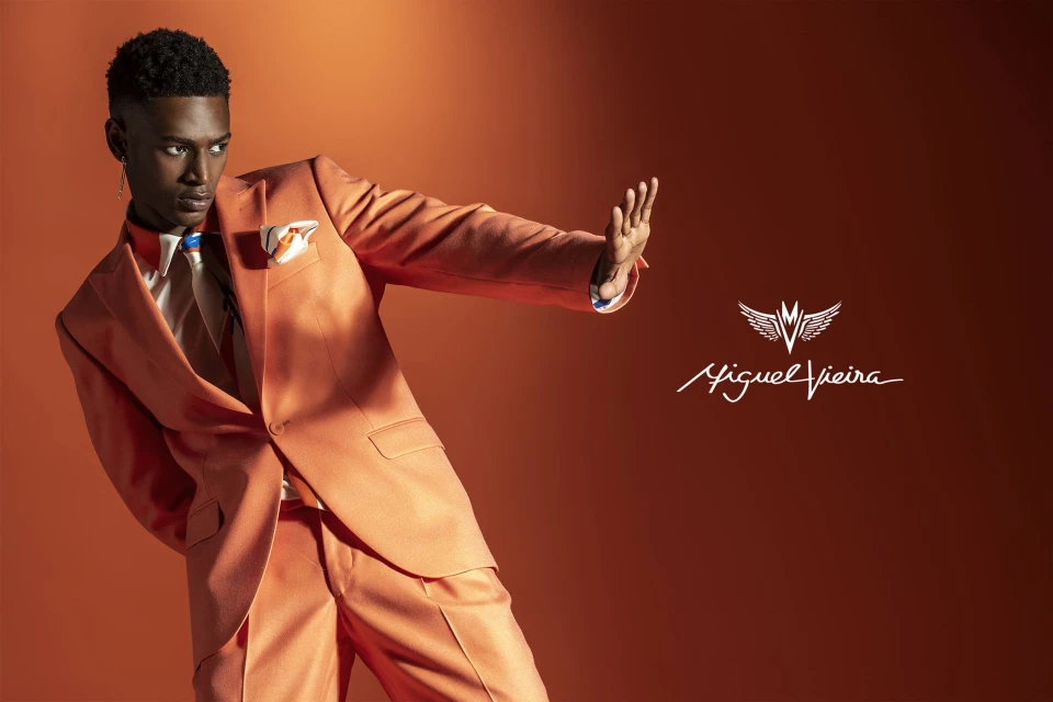 Stylish Black male model in a peach suit poses against a matching backdrop, showcasing modern fashion in a studio setting with logo.