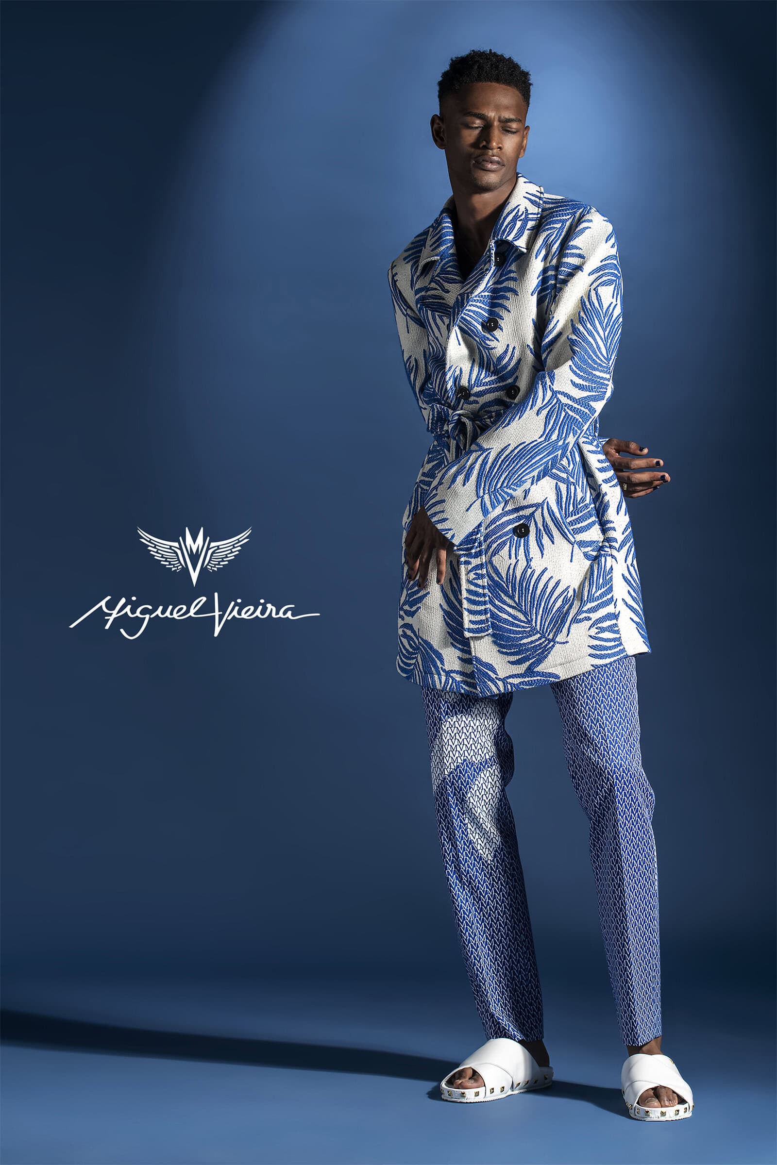 A stylish adult male model poses in blue and white patterned jacket and pants in a studio with moody lighting and the Miguel Vieira logo.