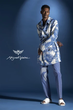 A stylish adult male model poses in blue and white patterned jacket and pants in a studio with moody lighting and the Miguel Vieira logo.