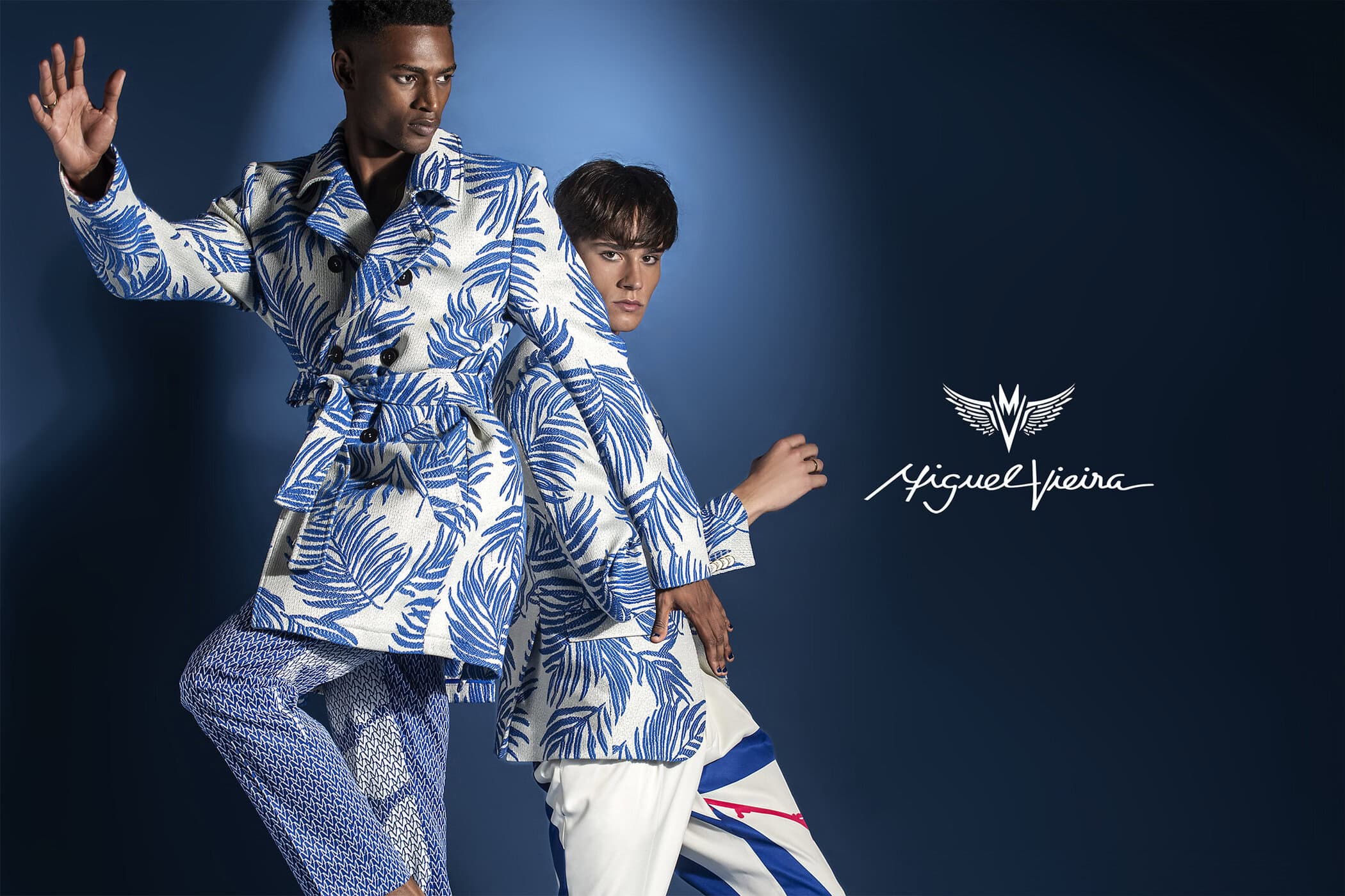 Two fashion models strike a pose in patterned trench coats and pants against a blue backdrop, promoting the Miguel Vieira brand.