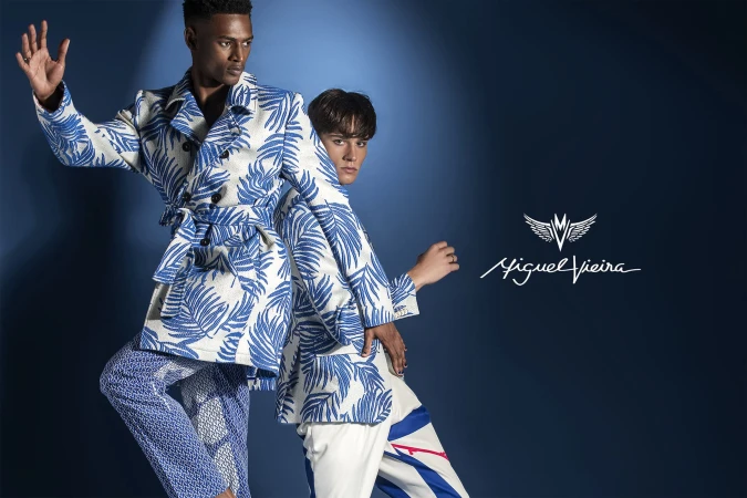 Two fashion models strike a pose in patterned trench coats and pants against a blue backdrop, promoting the Miguel Vieira brand.